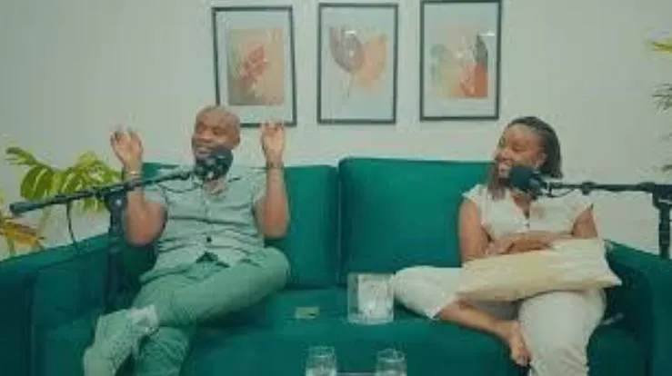 Two hosts laughing on a green sofa in front of podcast microphones